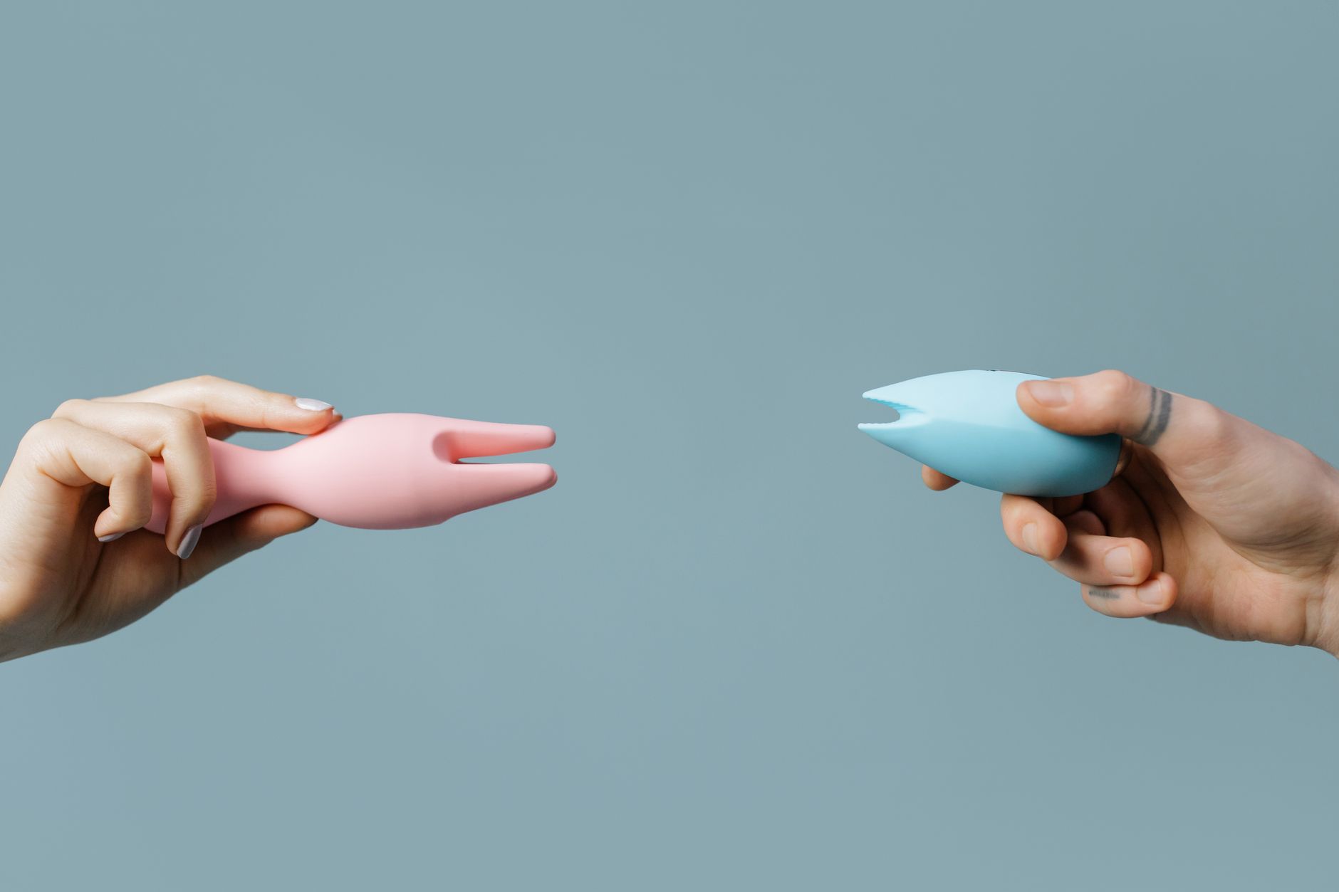 Two hands holding lemon-colored and blue silicone vibrators against a pastel background.